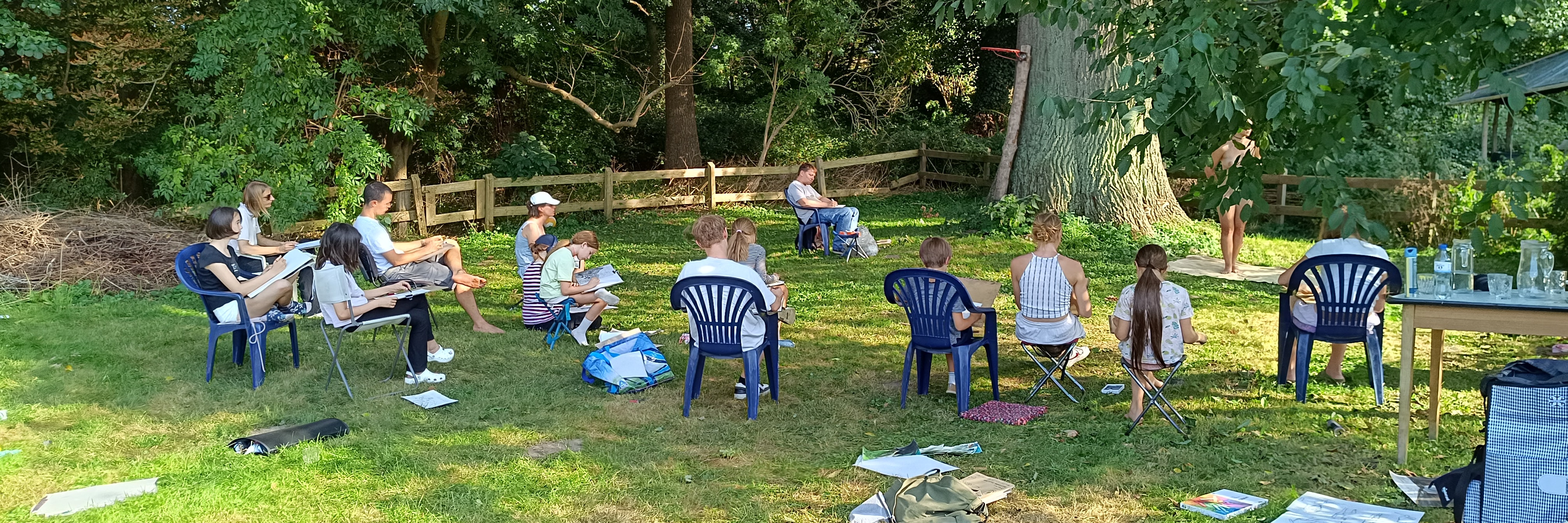 Gruppe von Erwachsenen und Kindern zeichnet im Freien eine stehende Person unter Bäumen.