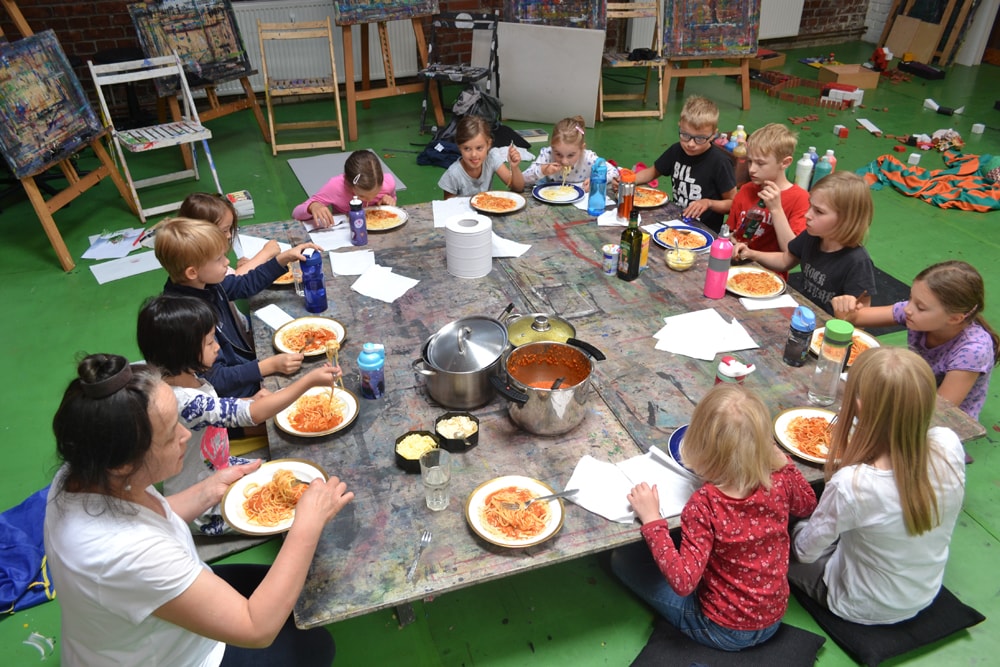 Kinder essen gemeinsam am großen Ateliertisch zu Mittag